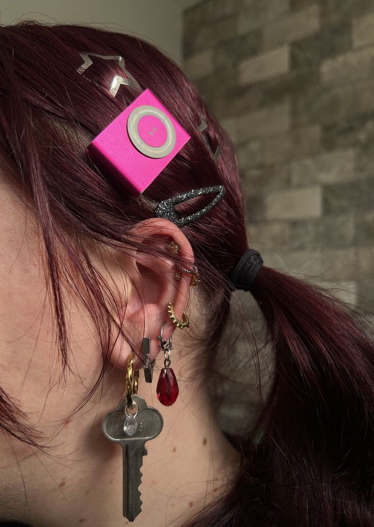 Person with dark red hair wearing a hot pink iPod Shuffle as a hair clip alongside multiple earrings.