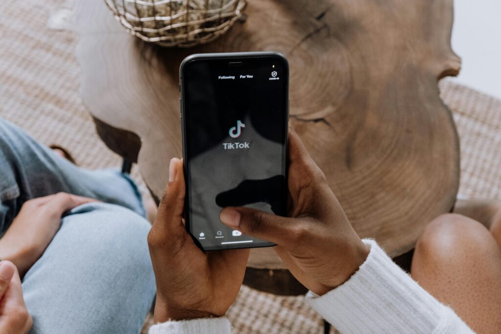 This image shows a person holding a smartphone, with their thumb about to press the TikTok app icon. The phone's screen displays a variety of other apps, including Instagram, Facebook, PayPal, and a messaging app, as well as some common system icons like the camera and settings. The person holding the phone appears to be sitting, and their hands are visible against the phone. The scene is indoors, with a light, casual atmosphere suggested by the person's relaxed hand positioning and clothing. The phone seems to be an iPhone based on its interface design and layout.