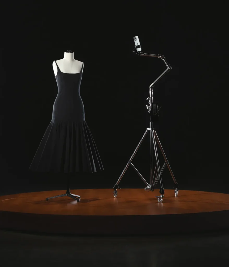 The image showcases a minimalist setup from the Jacquemus x Apple collaboration. A sleek black dress is displayed on a mannequin atop a round wooden platform, accompanied by a metallic tripod holding an iPhone. The dark background emphasizes the fusion of fashion and technology, highlighting the innovative partnership between Jacquemus and Apple.