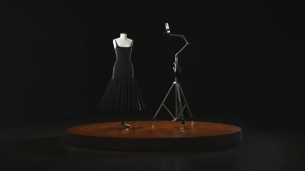 The image showcases a minimalist setup from the Jacquemus x Apple collaboration. A sleek black dress is displayed on a mannequin atop a round wooden platform, accompanied by a metallic tripod holding an iPhone. The dark background emphasizes the fusion of fashion and technology, highlighting the innovative partnership between Jacquemus and Apple.