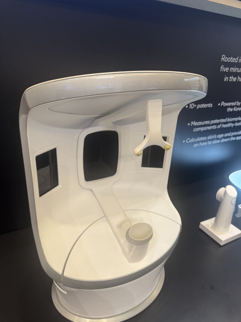 CES 2025 stand showing a device from L'Oréal, the Cell BioPrint: A futuristic white skincare analysis station with a curved design and built-in display areas. This station is used to collect and assess skin protein samples. It has a distinct hanging arm attachment and a flat surface for sample preparation. The setup highlights innovation in personalized skincare solutions.