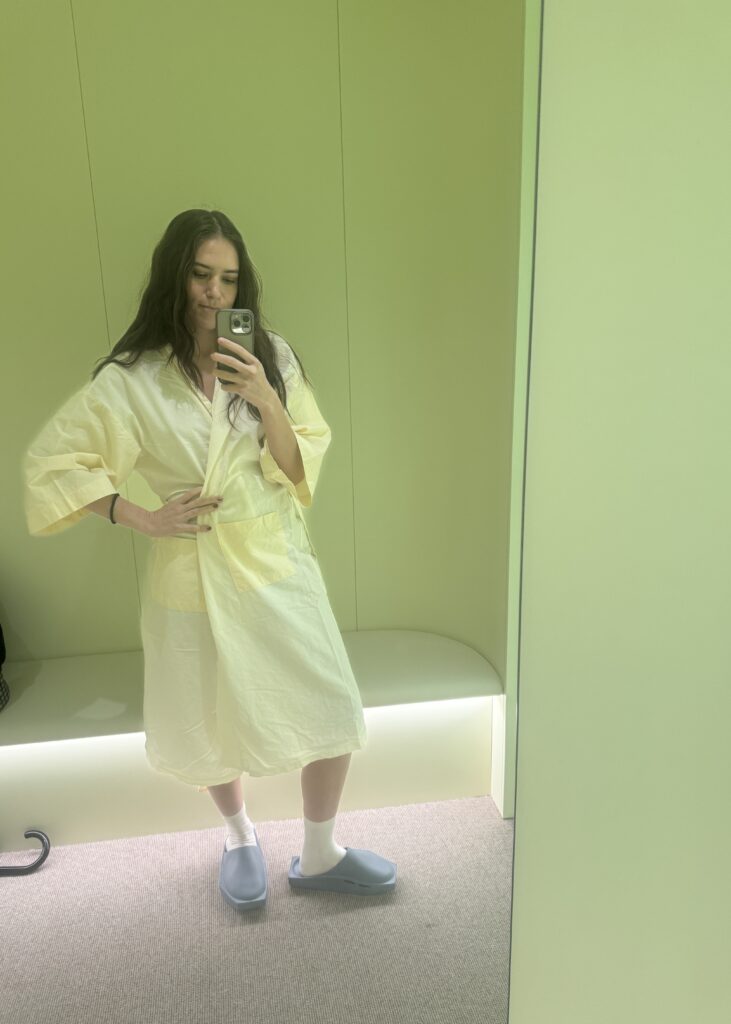 Tech journalist Natalie Salmon stands in a mirror selfie wearing Neko Health’s signature yellow robe and grey slippers, emphasizing comfort and casual technology.