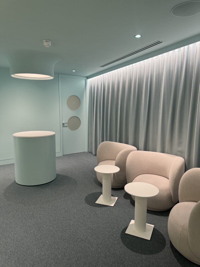 A calming teal room with modern, curved chairs and small white tables, offering a serene environment for Neko Health visitors, complemented by soft curtains and minimalist design elements.