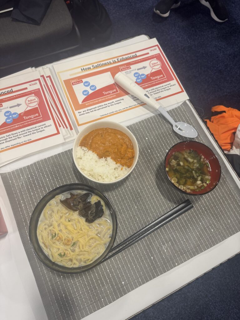 A display table featuring the Kirin Electric Salt Spoon alongside bowls of food like ramen, curry, and soup. Informative charts explain how the spoon works, using mild electrical currents to enhance saltiness on the tongue. The setup showcases the device’s innovative approach to low-sodium dining.