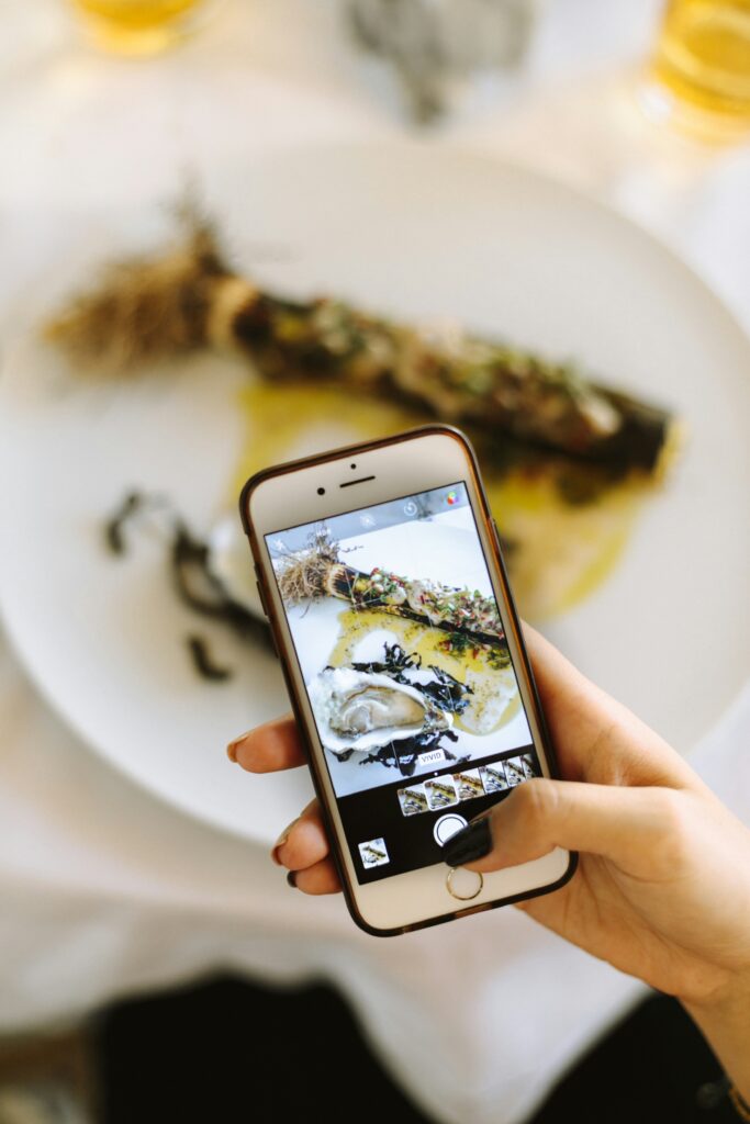 This image captures a person photographing a gourmet dish with a smartphone. The plate features a beautifully plated seafood dish, with vivid details of oysters and garnishes displayed on the phone screen, highlighting food photography's creative and social appeal.