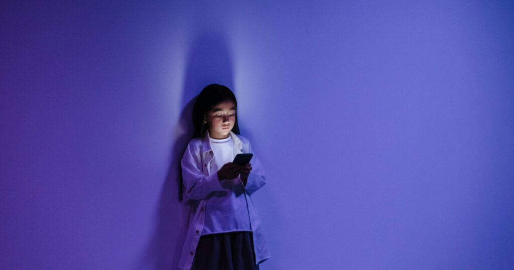 A young girl stands against a purple wall at night, illuminated by the blue light from her smartphone screen, symbolising children’s relationship with technology.