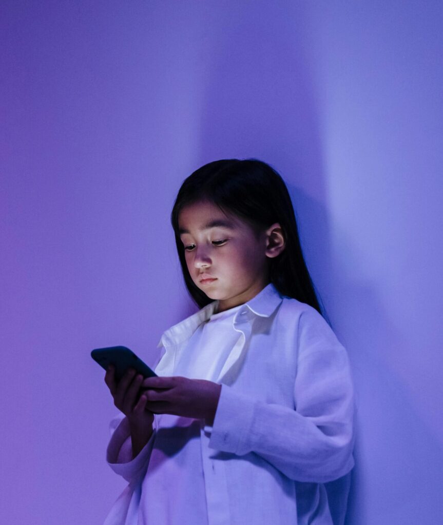 A girl in a white shirt looks down at her smartphone under soft purple lighting, representing the impact of mobile devices on childhood and attention.