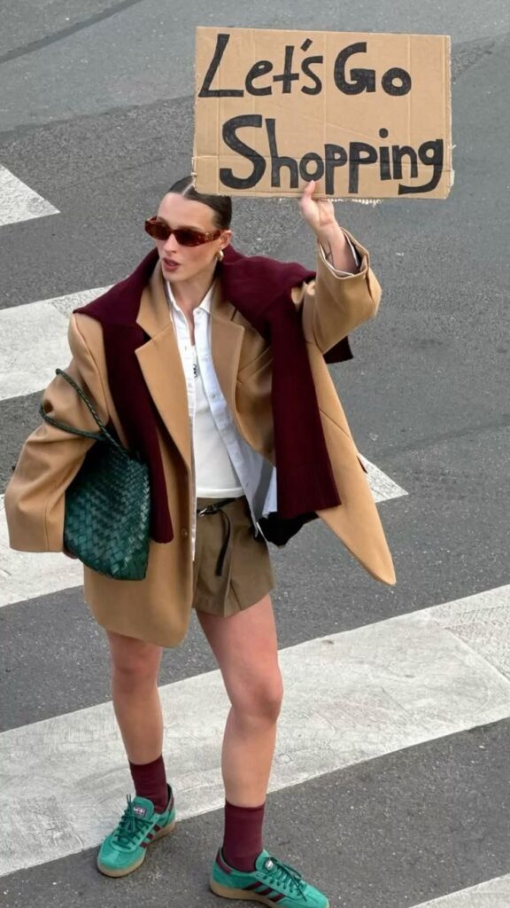 Stylish woman in oversized blazer, sneakers, and sunglasses holding a cardboard sign reading “Let’s Go Shopping” on a city street.