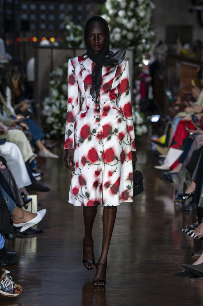 Model wearing a white coat dress with red rose floral print, crystal buttons, and a black sheer headscarf from Patrick McDowell’s Spring Summer 2026 collection at London Fashion Week.