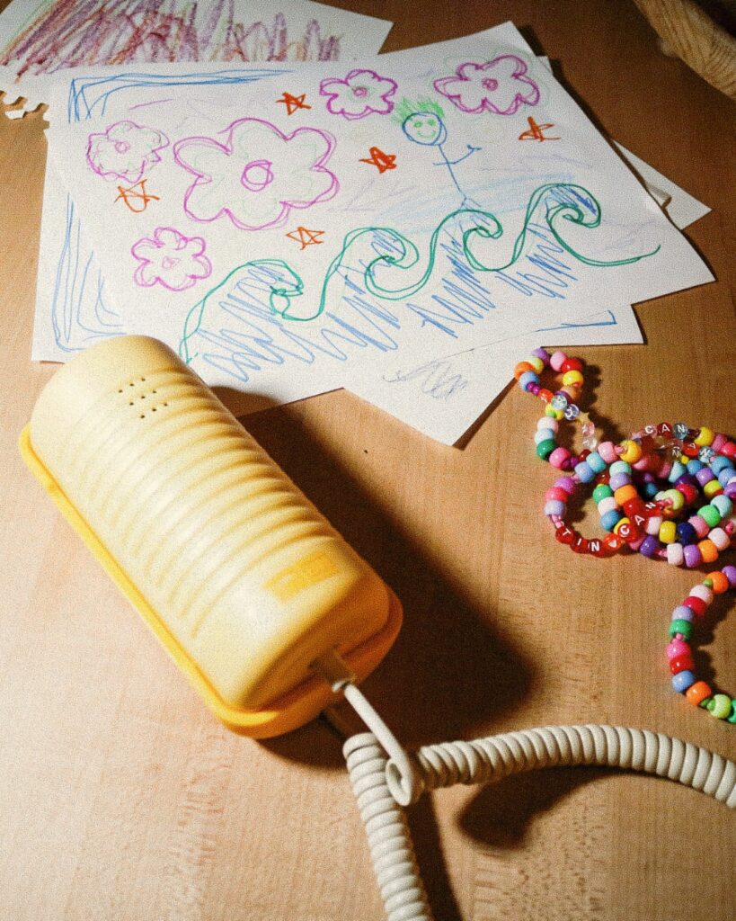 A yellow Tin Can phone rests on a wooden desk surrounded by colourful children’s drawings and beaded bracelets.