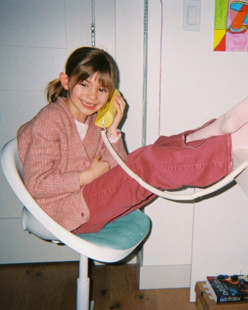 A smiling child sits in a chair holding a yellow Tin Can phone to her ear, dressed in a pink sweater and trousers.