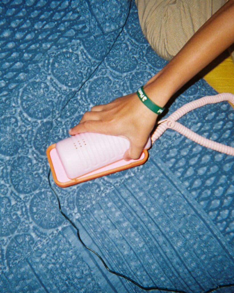 A hand grips a pink Tin Can phone against a patterned blue blanket, the coiled cord trailing across the frame.