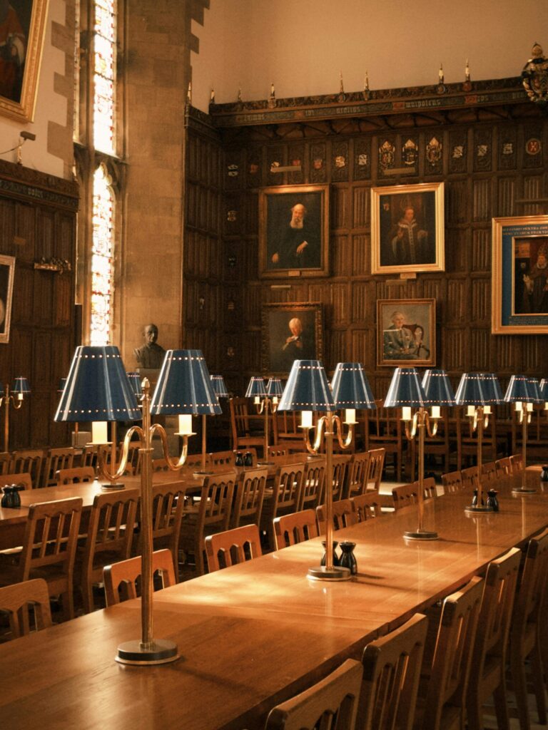 Interior of an Oxford University dining hall with long wooden tables, blue-shaded lamps, and historic portraits on wood-paneled walls.
