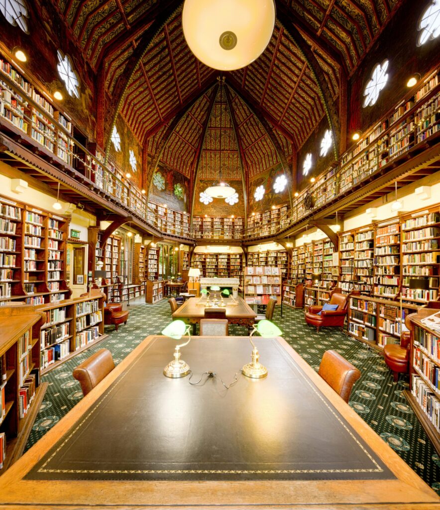 Interior of a historic Oxford library with vaulted wooden ceilings, tall bookshelves, and green desk lamps.