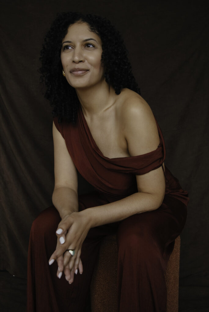 Portrait of Phoebe Parke in a burgundy one-shoulder dress, seated and smiling slightly against a brown backdrop, photographed by Krystal Neuvill.