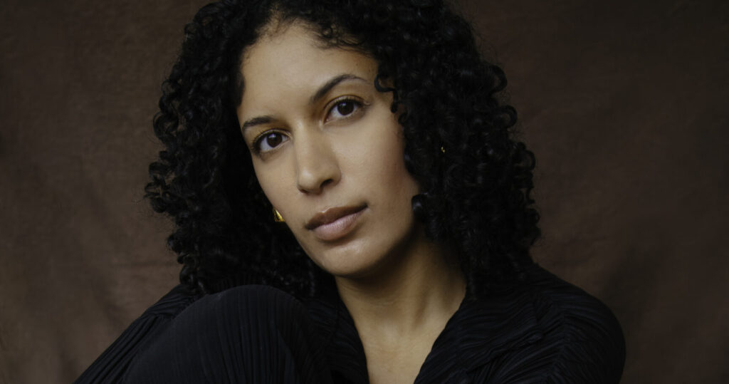 Portrait of Phoebe Parke wearing a black pleated outfit, seated on an orange chair with a thoughtful expression against a brown backdrop, photographed by Krystal Neuvill.