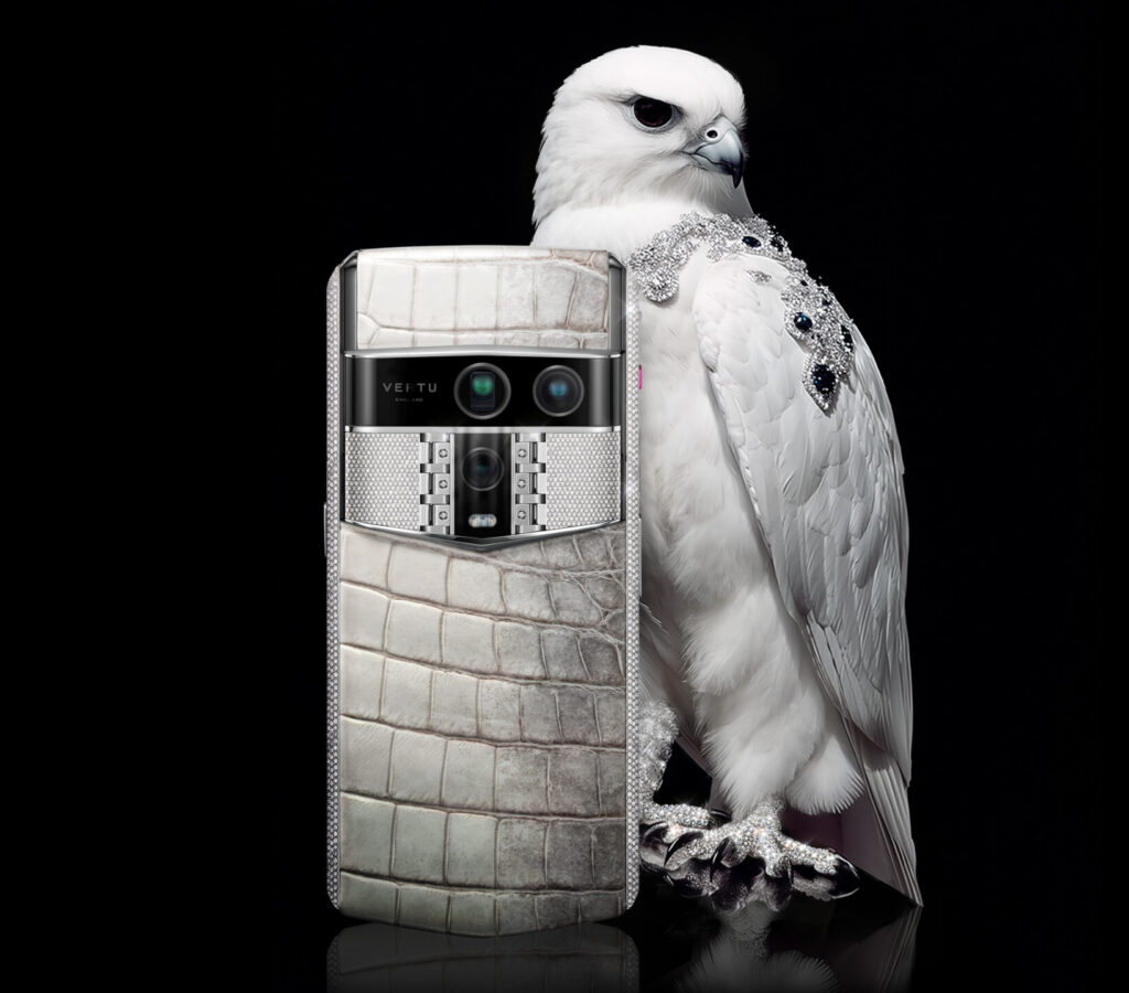 Vertu smartphone in white crocodile leather finish displayed beside a white falcon adorned with diamond jewelry, representing elegance and craftsmanship.