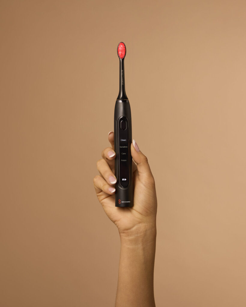 Close-up of a person holding the BON CHARGE Red Light Toothbrush upright against a beige background, highlighting its minimalist black finish and LED bristles.