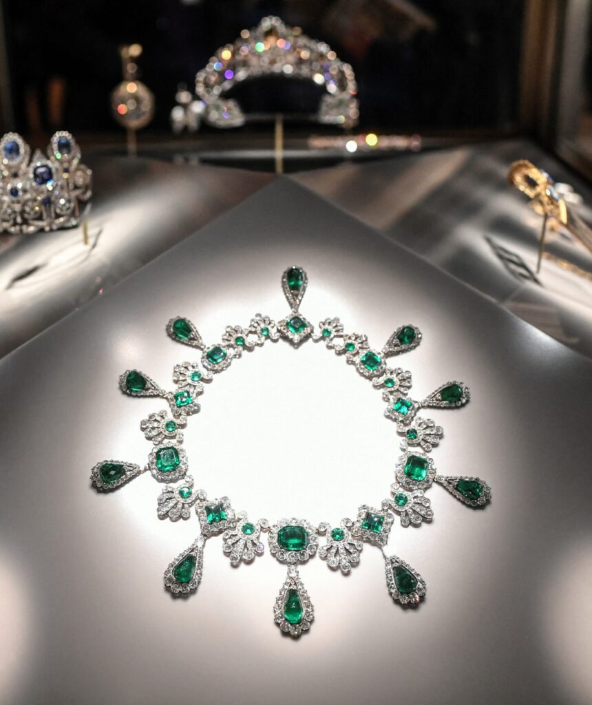 A close-up photograph of diamond and emerald royal jewellery displayed at the Louvre Museum in Paris, including a necklace and earrings once belonging to Empress Eugénie.