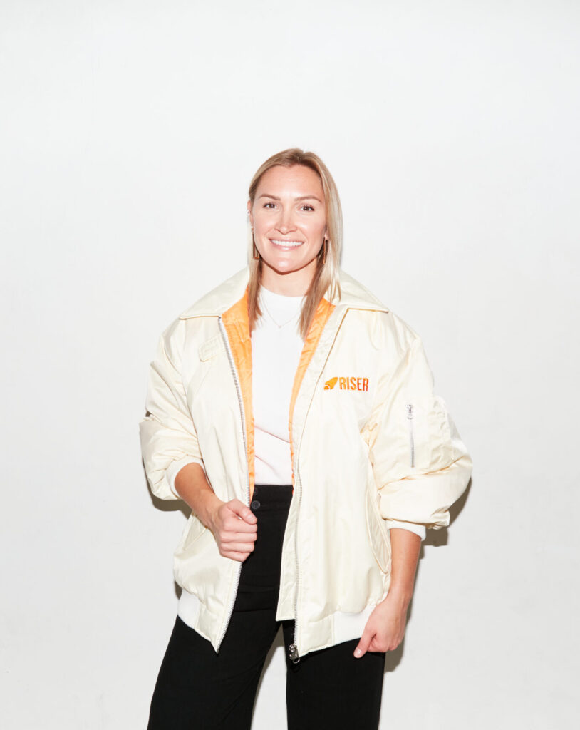 Suz Bannister, co-founder of RISER, poses against a white backdrop wearing a cream RISER-branded jacket and smiling confidently.