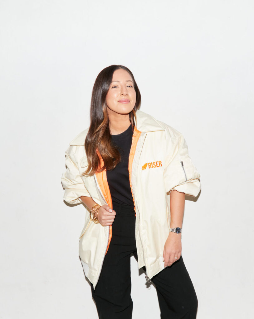 Lamees Butt, co-founder of RISER, stands against a white background wearing a cream RISER jacket with orange lining, hands in pockets and smiling.
