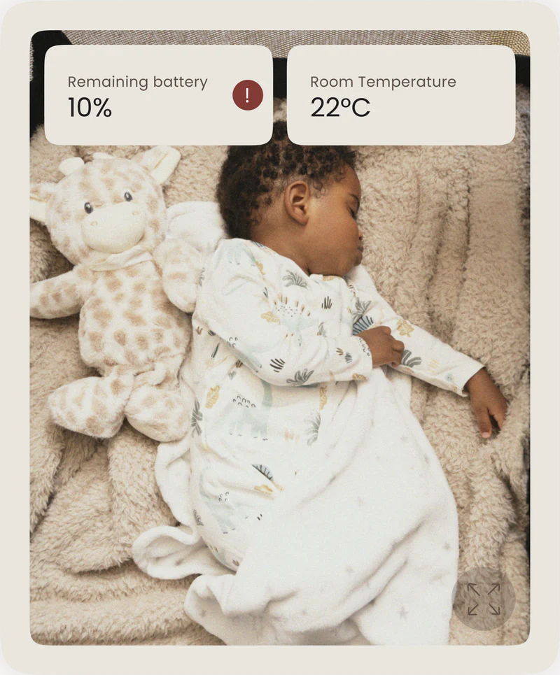Sleeping baby beside a stuffed giraffe toy with on-screen Dote app notifications showing room temperature and battery status.