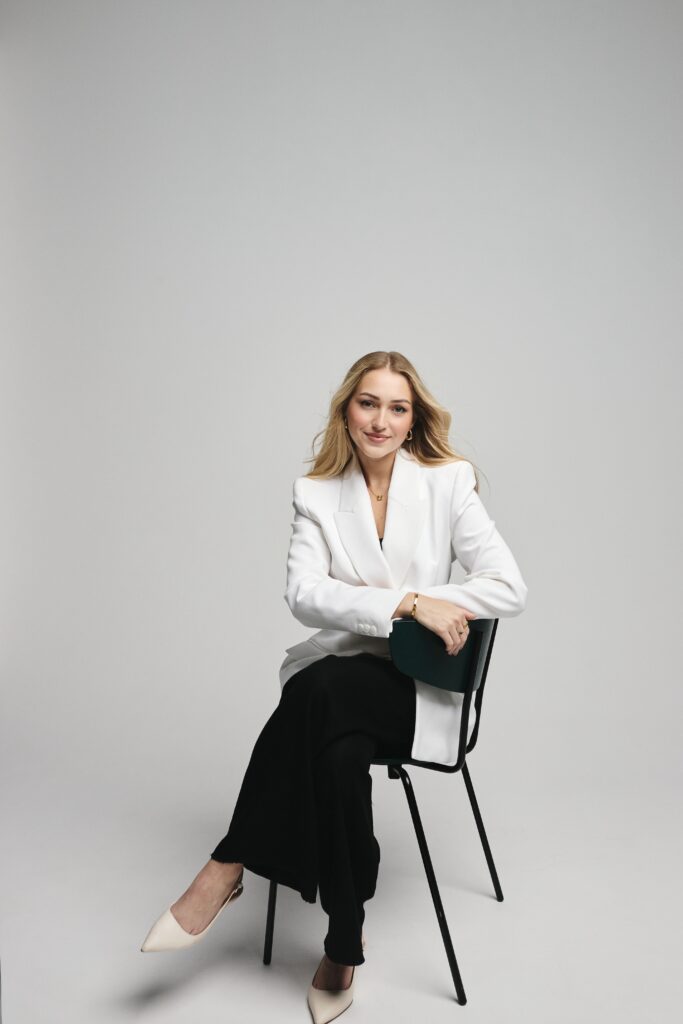 Nicola Gunby, Co-Founder and CMO of CLIQ, seated on a black chair wearing a white blazer and black trousers in a minimalist studio portrait, representing the new era of social networking and tech entrepreneurship.