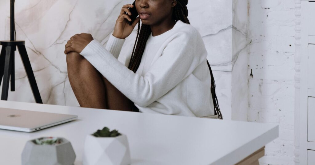 Woman sitting at a white desk in a bright modern home office, speaking on the phone with a laptop nearby, illustrating secure remote work practices.