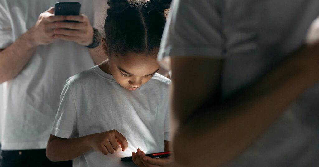 A young girl with braided buns looks intently at her smartphone while surrounded by adults in white shirts, symbolising generational screen dependence.