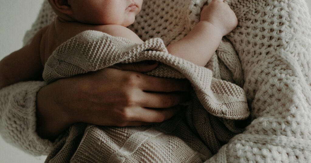Close-up of a baby wrapped in a beige knitted blanket being held by a parent wearing a cream chunky sweater.