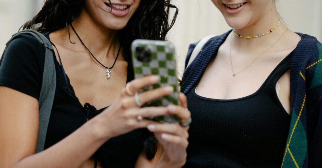 Two young women smiling while looking at a smartphone outdoors, representing influencers engaging with social media content.