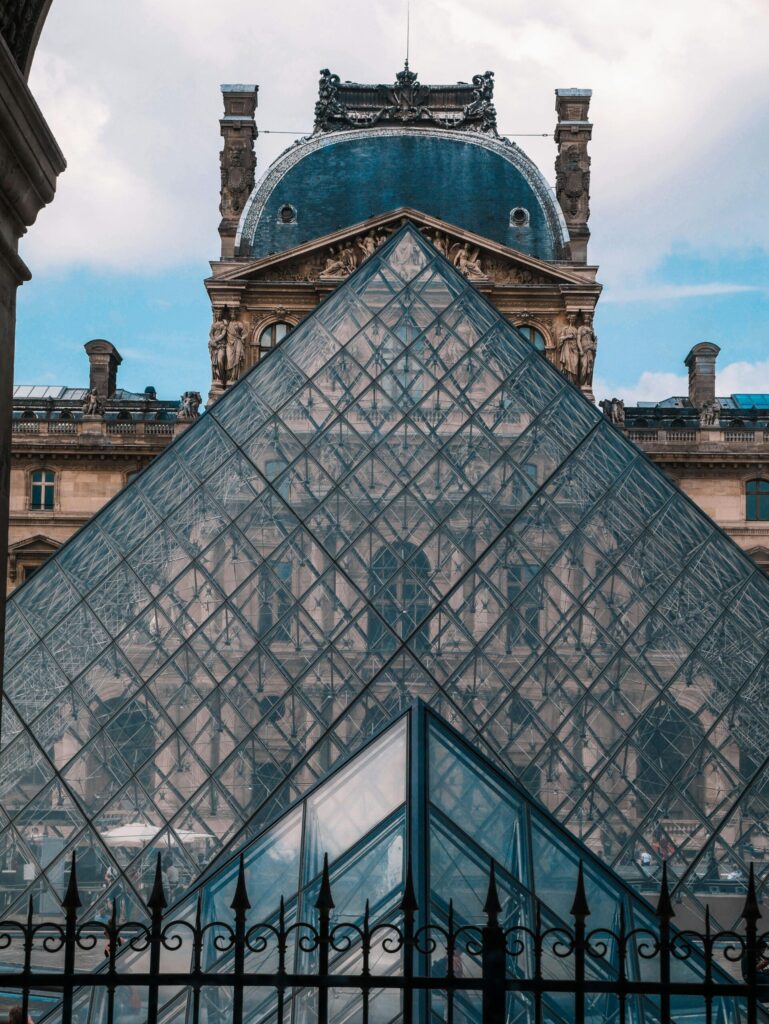 The glass pyramid of the Louvre Museum in Paris reflecting the historic dome and façade of the palace on a bright day.