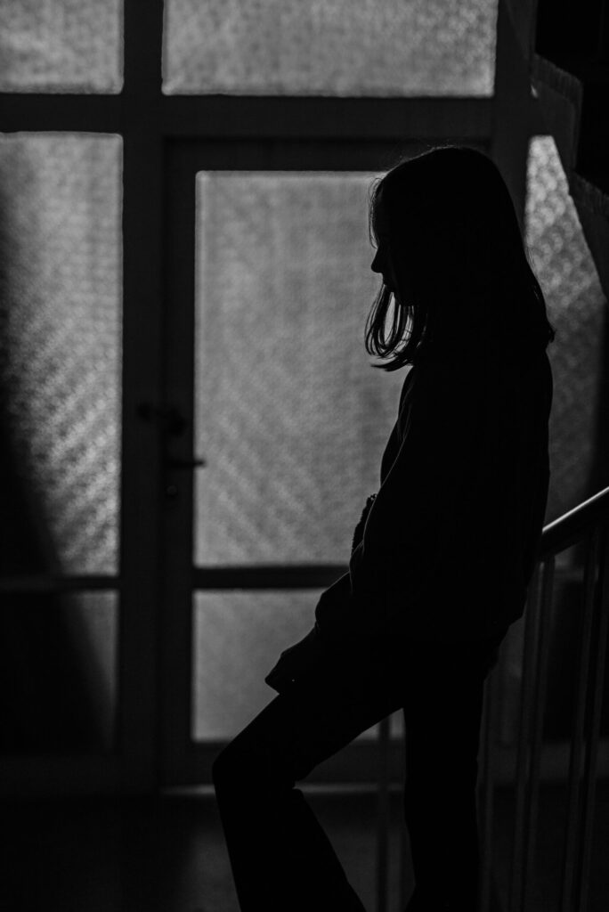 Black and white photo of a young girl standing in silhouette near a window, symbolising online vulnerability, child safety, and the hidden impact of digital abuse in the age of social media.