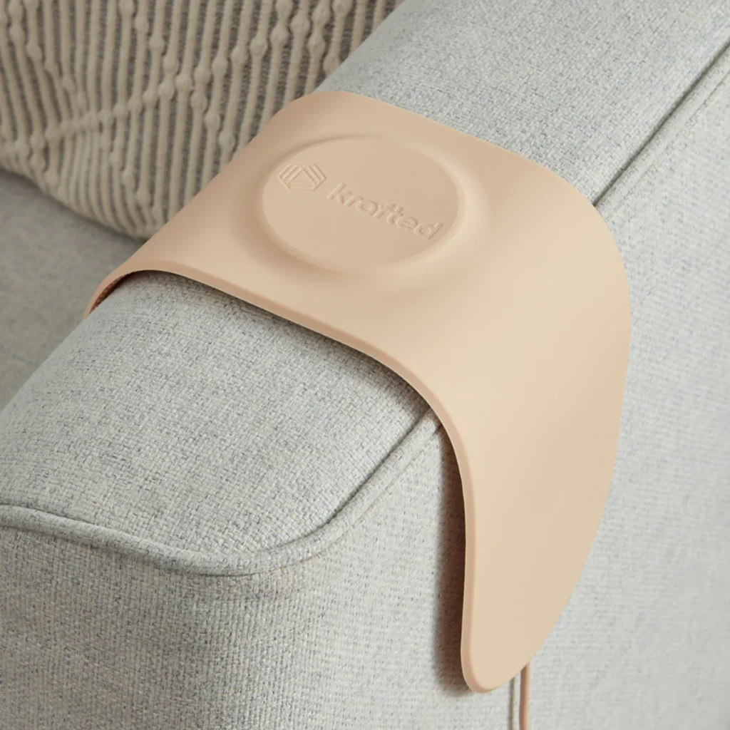 A beige Krafted wireless charging pad draped over a light grey sofa armrest, designed to charge devices hands-free while blending into home décor.