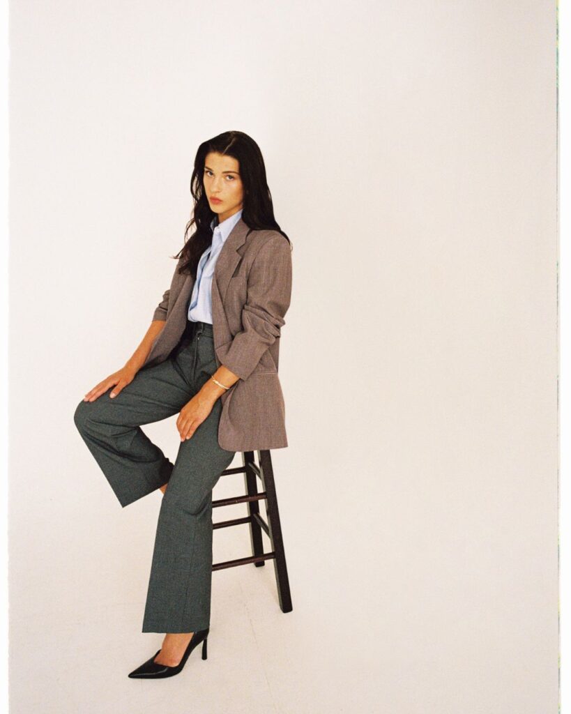 Phoebe Gates sits on a stool in a neutral studio setting wearing tailored trousers, a light blue shirt, and a grey blazer.