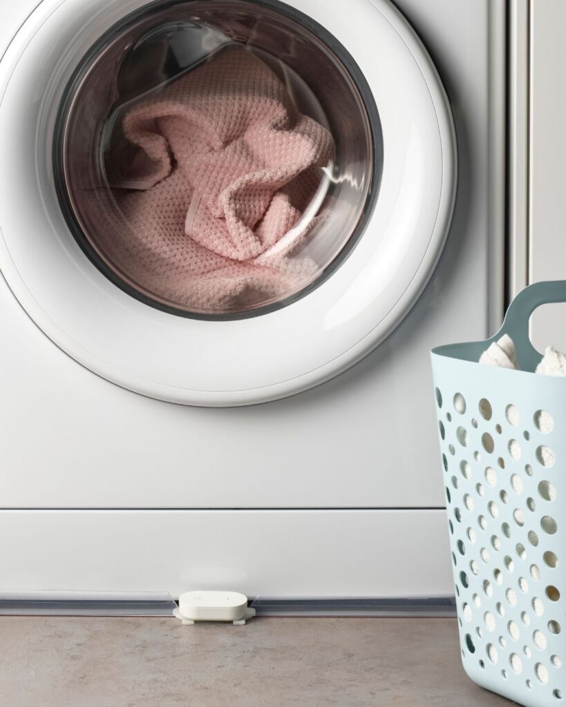IKEA KLIPPBOK smart water leakage sensor placed under a washing machine with a pink towel inside the drum.