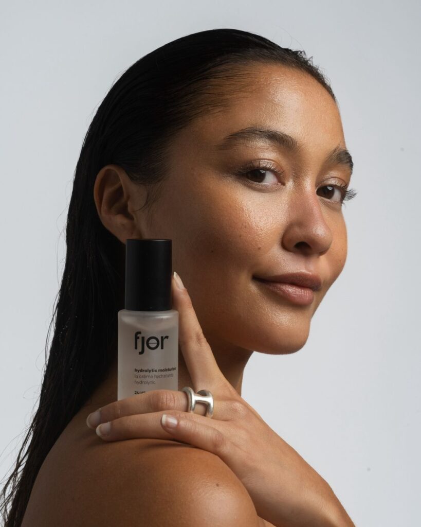 Natalie Enslöw holding a bottle of fjör Hydrolytic Moisturiser, with glowing skin and sleek hair against a white backdrop.