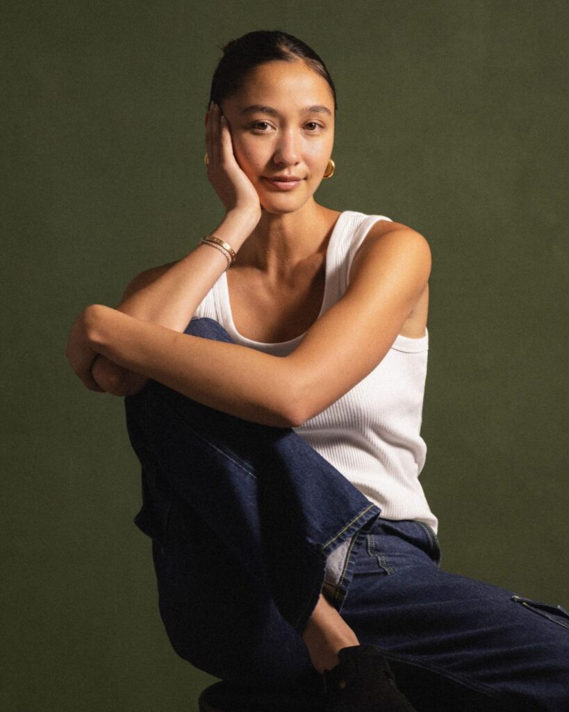 Natalie Enslöw seated against a green background in a white tank top and gold jewellery, exuding natural confidence.