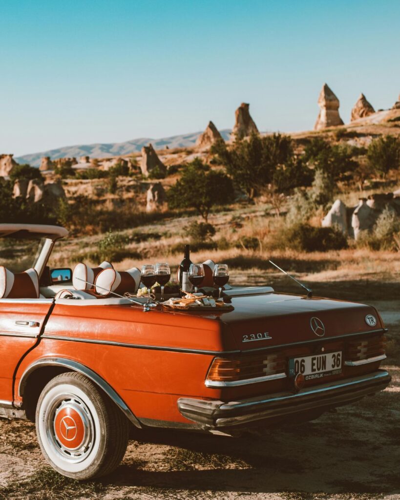 Red vintage Mercedes convertible with a picnic setup of wine and glasses in a scenic countryside landscape at sunset.