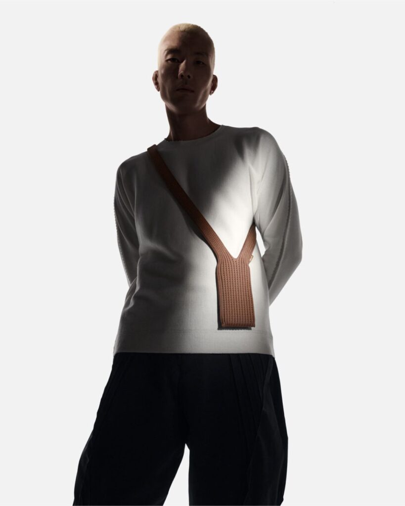 Model wearing a brown Issey Miyake iPhone Pocket crossbody accessory over a light sweater, standing in soft studio lighting.