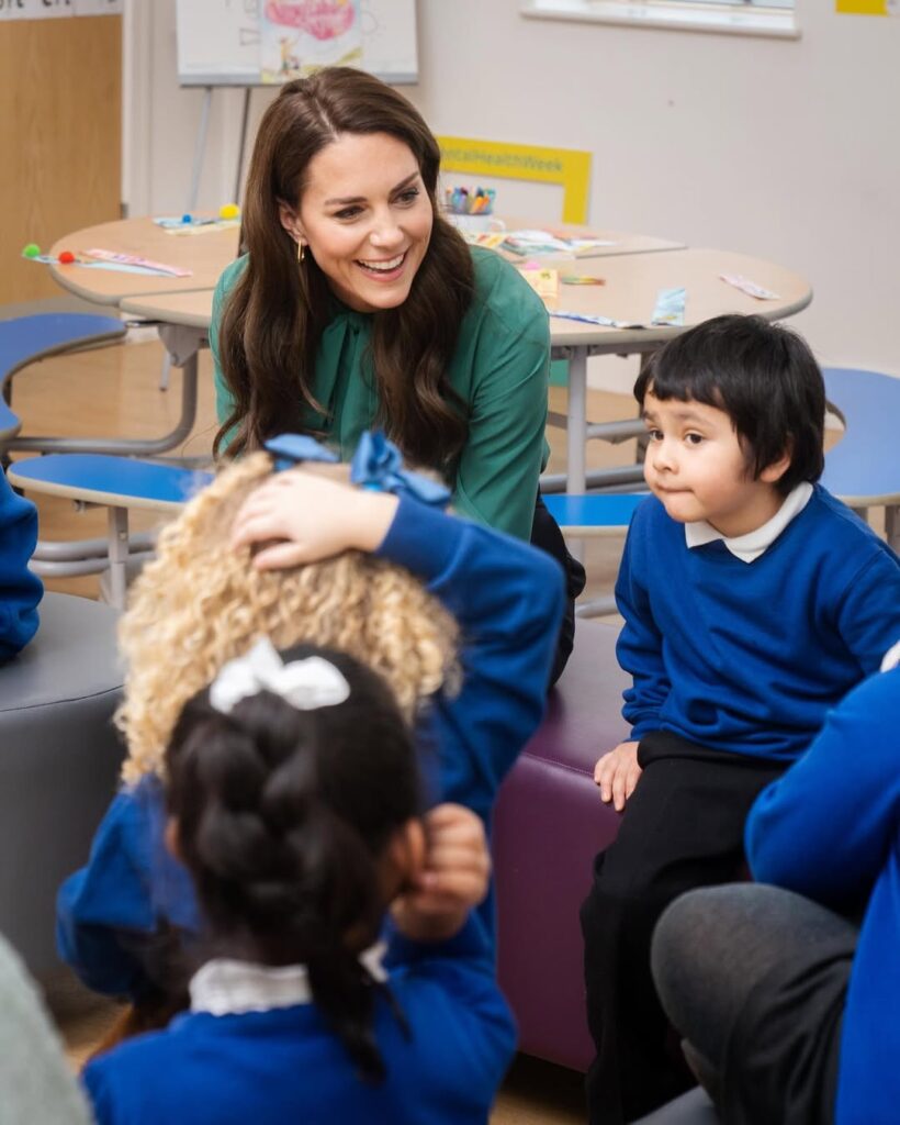 Princess Kate wearing a green blouse, sitting with young schoolchildren in a classroom setting and smiling during an early childhood engagement.