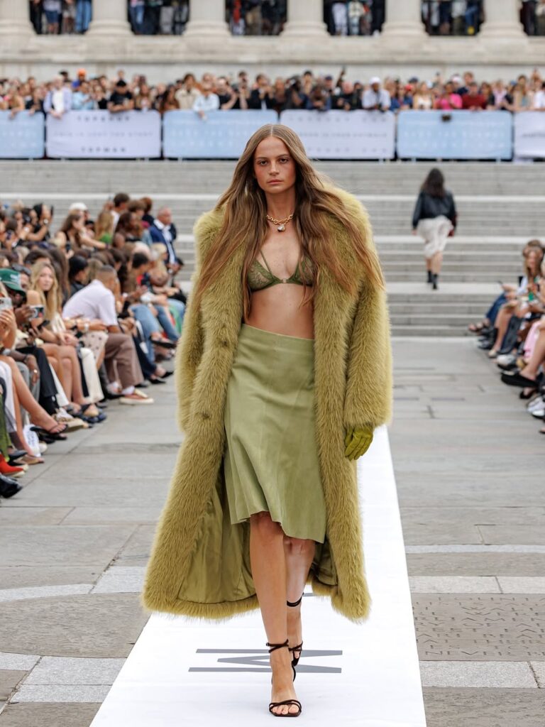 Model walking the Topshop SS25 runway in a long olive faux-fur coat, green bralette, and matching satin midi skirt.