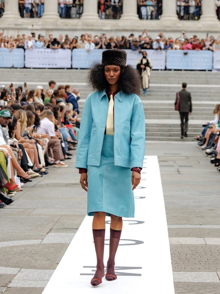 Model walking the Topshop SS25 runway in an aqua jacket and matching skirt, styled with sheer burgundy socks and a textured velvet hat during London Fashion Week.