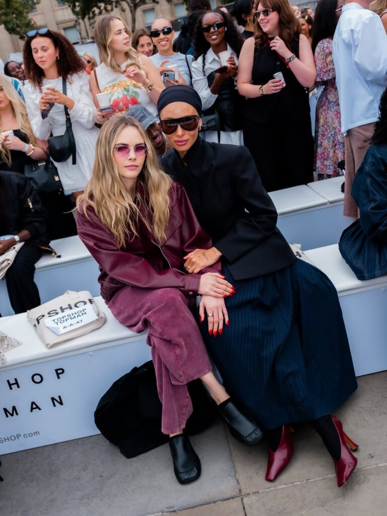 Cara Delevingne sitting front row at the Topshop SS25 show wearing oxblood corduroy, posing with Adwoa Aboah dressed in dark tailoring and sunglasses.