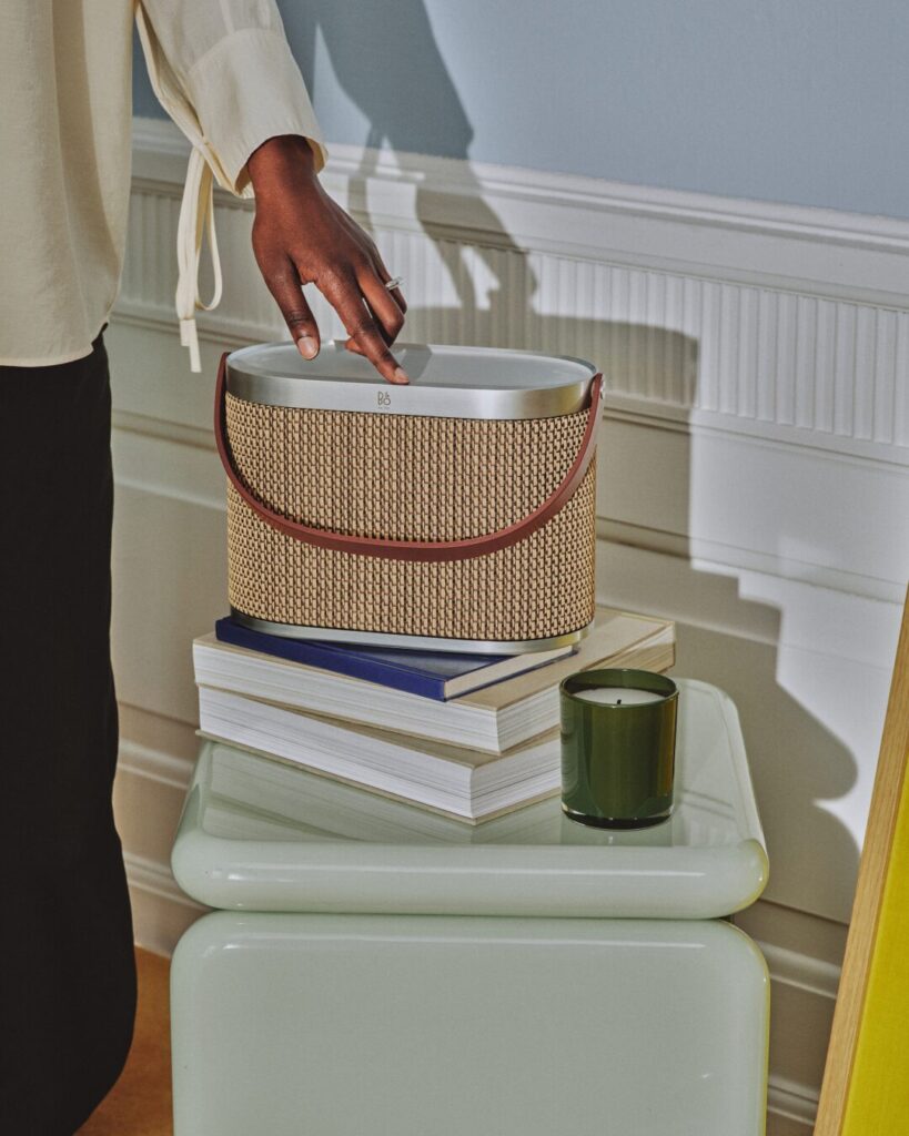 A hand reaches toward the Bang & Olufsen Beosound A5 portable speaker in beige raffia weave with brown leather handle, styled on a stack of design books.