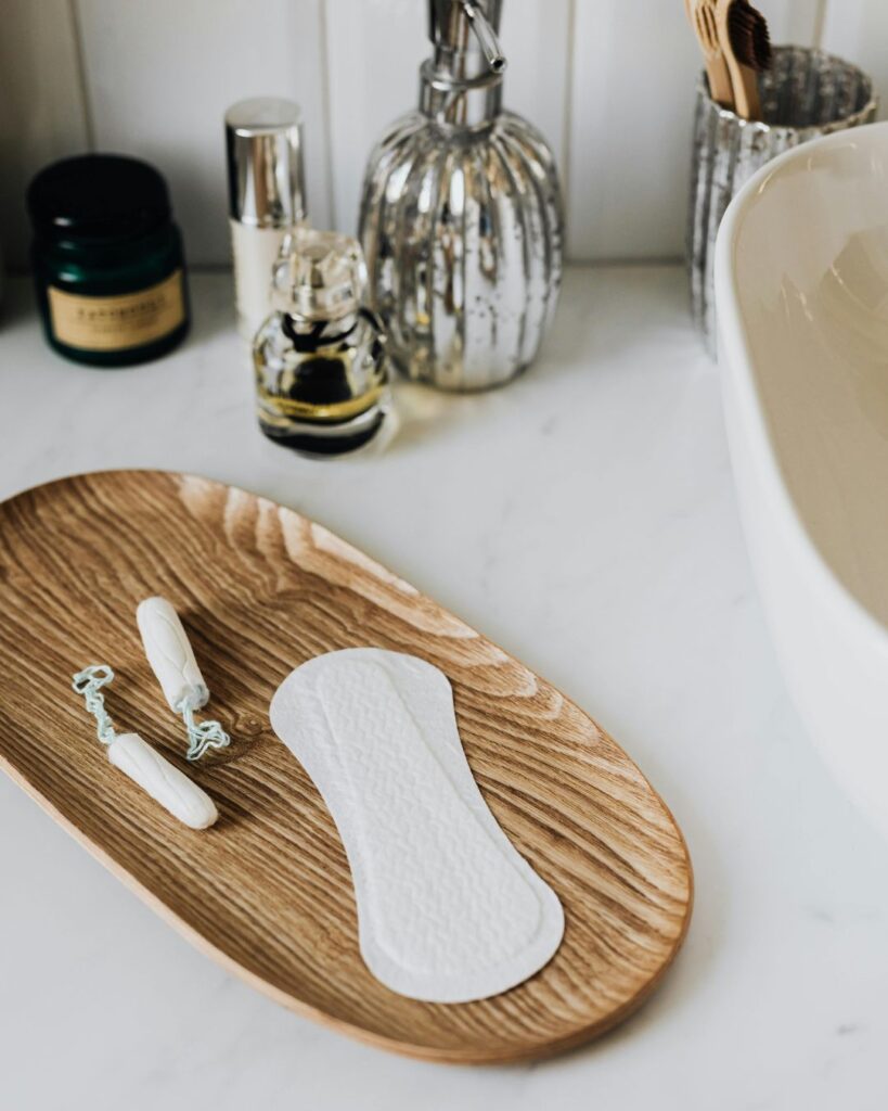 A minimalist wooden bathroom tray displaying tampons and a sanitary pad beside skincare products and a sink, symbolising modern menstrual health and hygiene awareness.