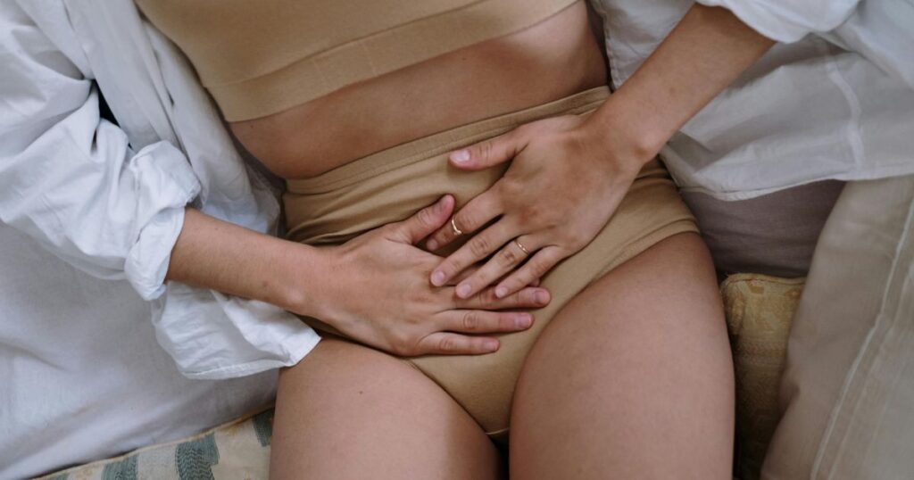 A woman in beige underwear and a white shirt holds her stomach while experiencing period pain, representing the ongoing challenges in women’s reproductive health and access to contraception.