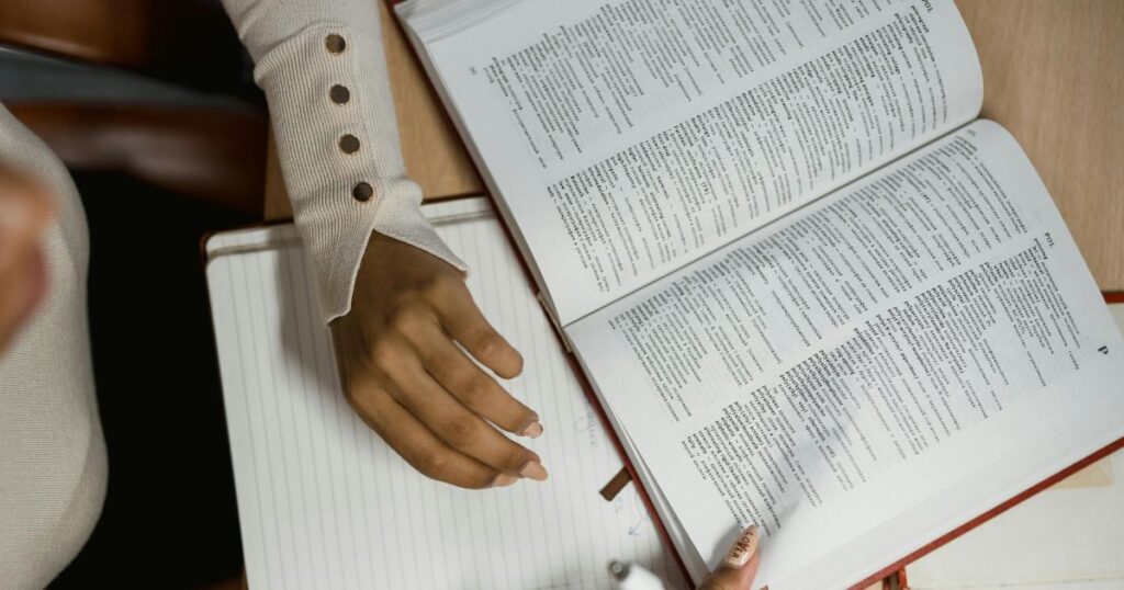 A person wearing a beige ribbed top studies an open dictionary on a desk with a notebook, symbolising learning, linguistics, and digital culture.
