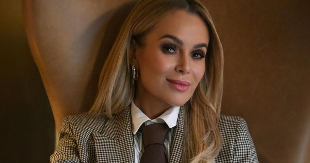A portrait of Amanda Holden wearing a checked blazer, white shirt and brown tie, posing indoors with soft lighting.