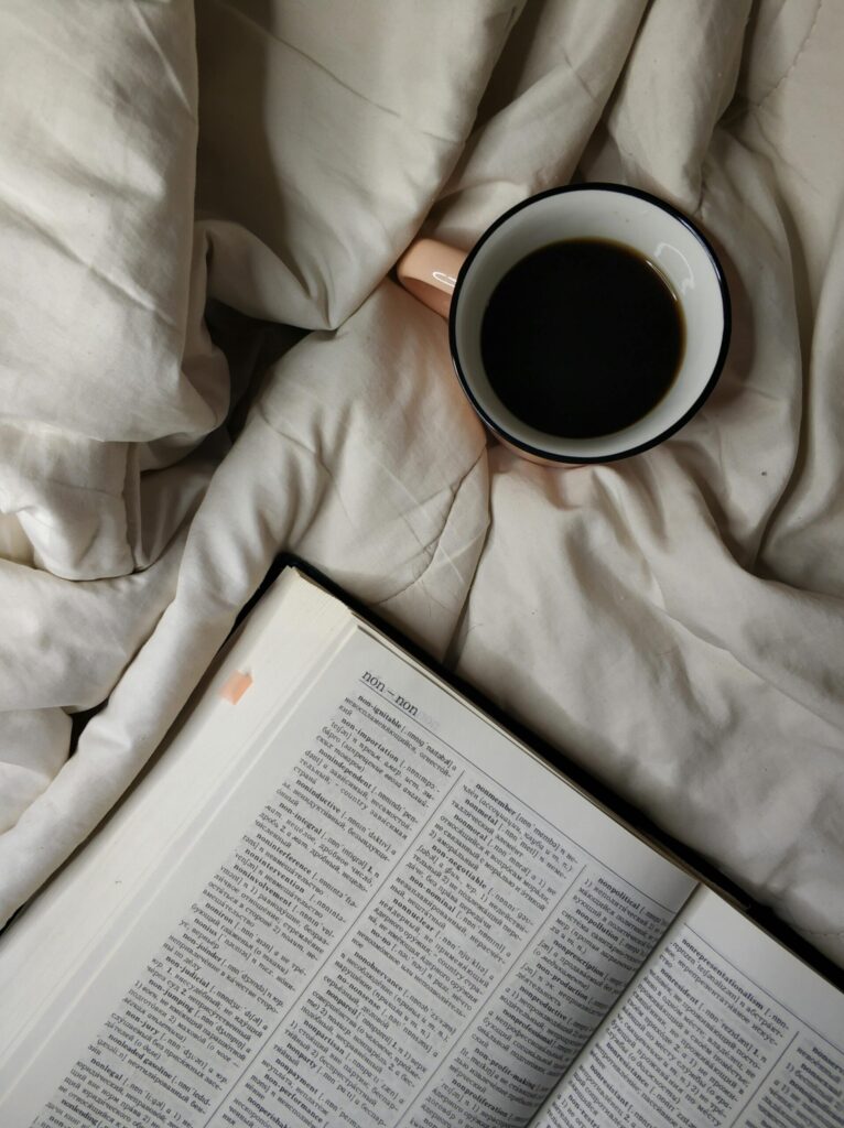 A cup of black coffee on white bedding beside an open dictionary, creating a cozy minimalist flatlay that evokes study, reading, and slow living.